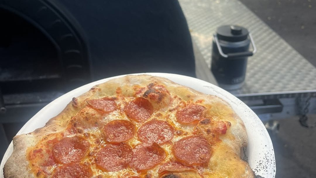A wood-fired pepperoni pizza on a plate in front of the Outrigger pizza oven and truck